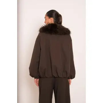 A model shown from the back wearing a dark brown jacket with a large dark brown faux-fur collar, paired with matching dark brown trousers, against a plain off-white background.