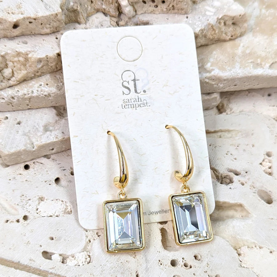 Gold earrings with square gemstones on a 'sarah tempest' branded card against a stone background.