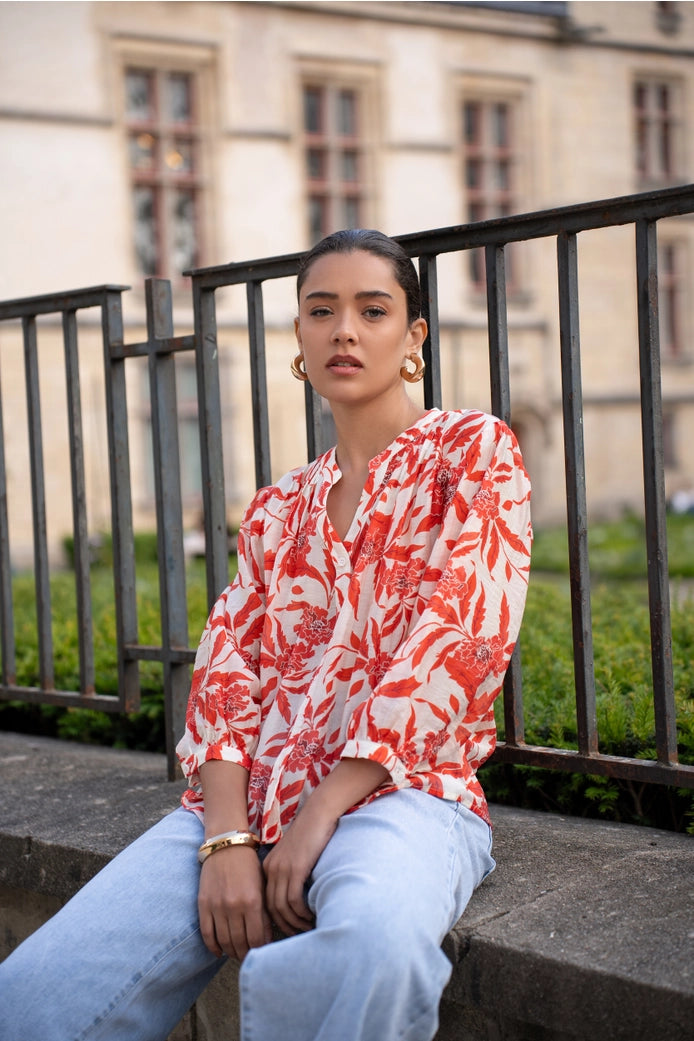 Woman wearing a red and white patterned blouse sitting on a ledge outdoors.