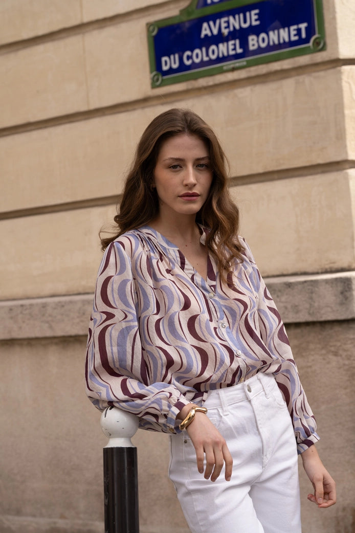 Woman in a patterned blouse and white pants standing against a beige wall with a street sign in the background.