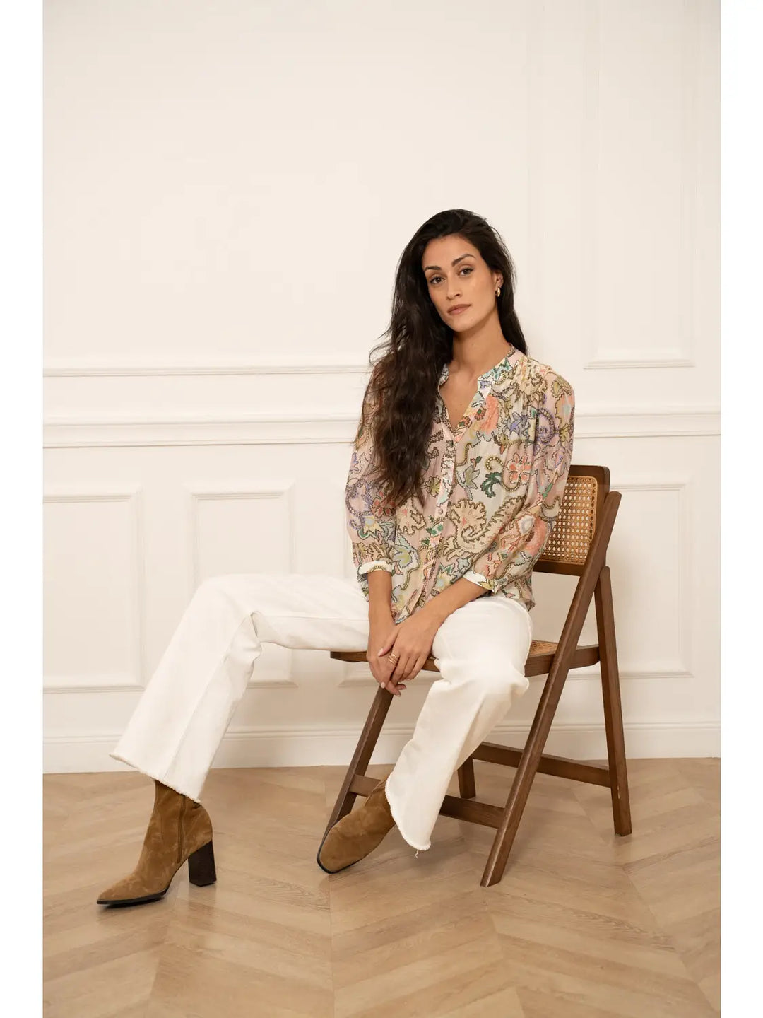 Woman sitting on a wooden chair wearing a floral blouse and white pants in a room with white walls.