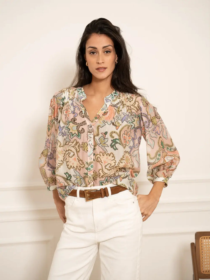 Woman wearing a patterned blouse and white pants against a plain background
