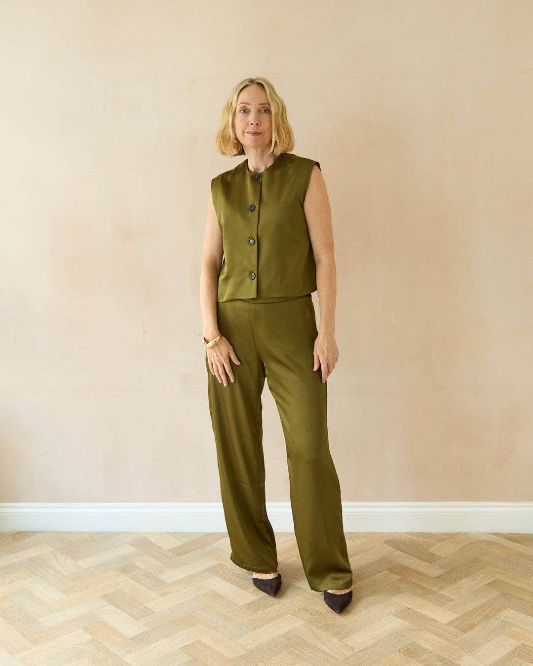 Woman wearing a green sleeveless top and pants standing against a beige wall.