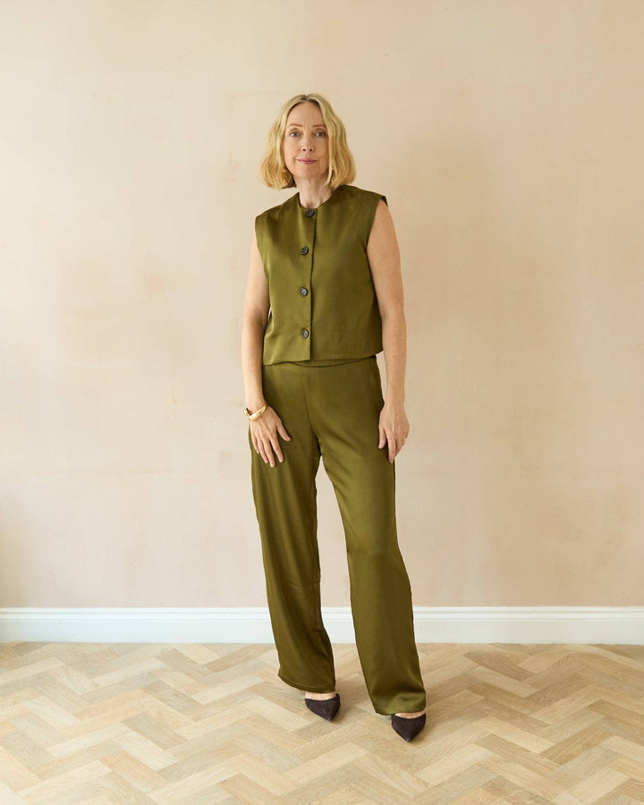 Woman wearing a green sleeveless top and pants standing against a beige wall.