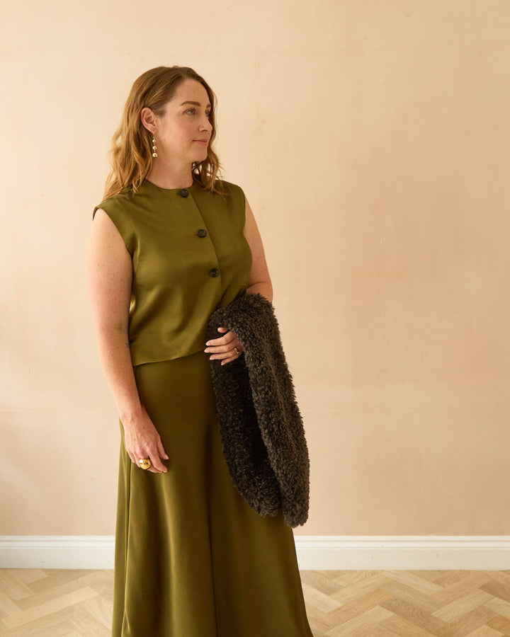 Woman wearing an olive green sleeveless top and skirt with a dark fur coat against a beige wall.