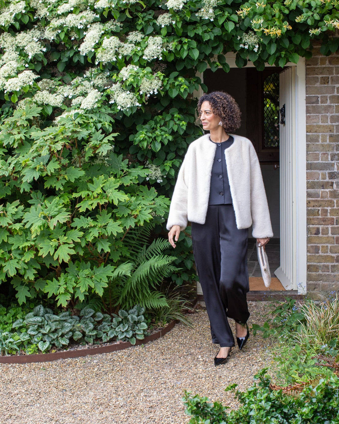 Woman walking outdoors in a garden setting with greenery and a building in the background