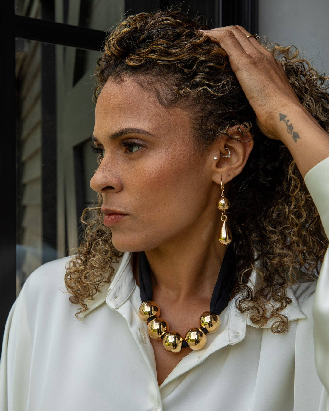 Woman wearing gold jewelry, including earrings and a necklace, with a blurred background.
