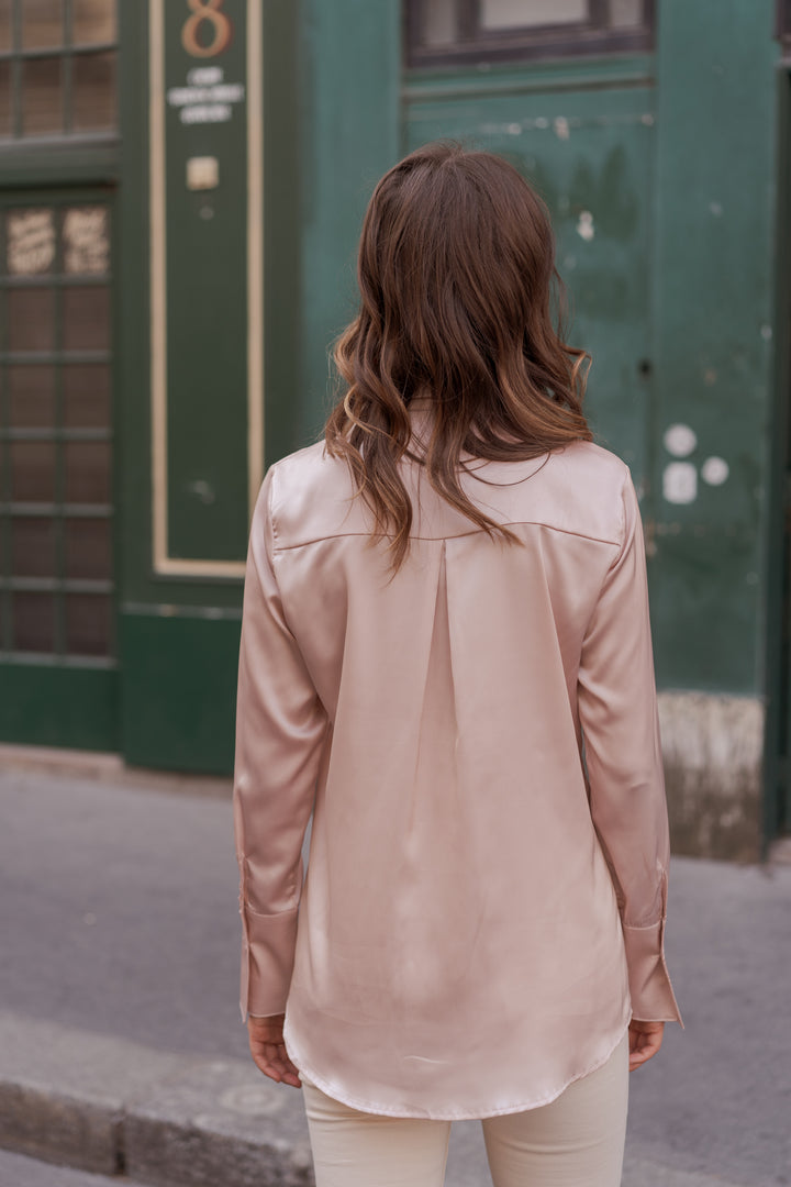 A woman viewed from behind wearing a satin-like blush pink long-sleeve blouse and light beige pants, standing outdoors in front of a dark green building.