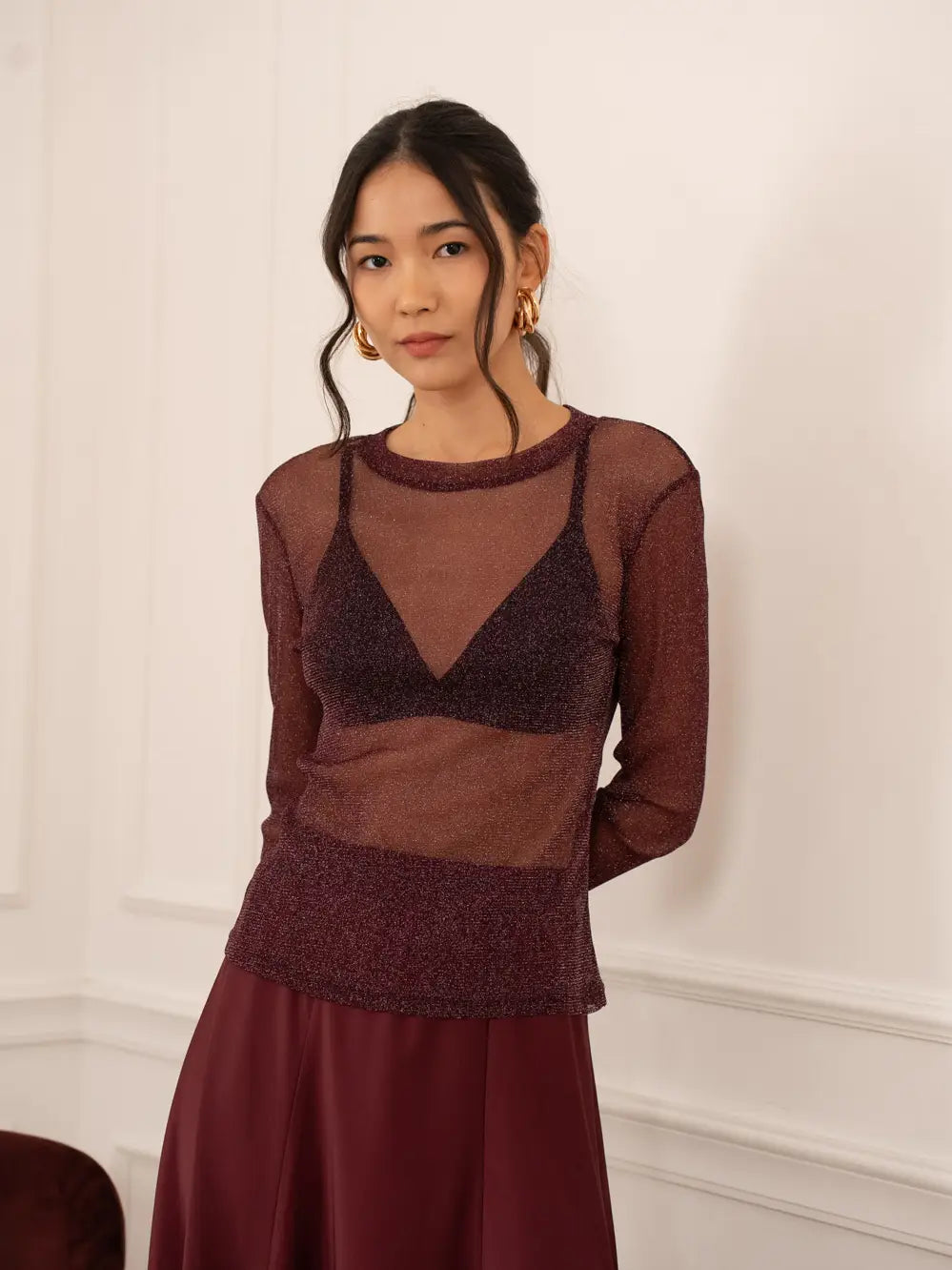 Woman wearing a sheer brown top and dark red skirt against a white wall.