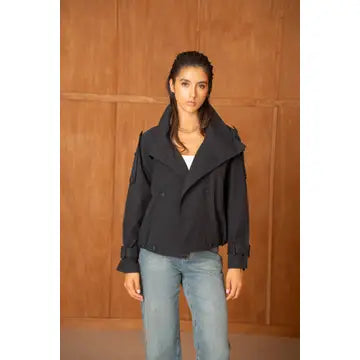 A woman modeling a black double-breasted jacket with a high collar, paired with light blue jeans, standing against a brown wooden paneled wall.
