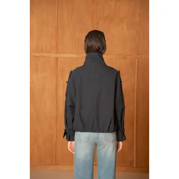 A person viewed from behind wearing a black long-sleeve jacket with shoulder epaulets and light-wash blue jeans, standing against a warm wood-paneled background.