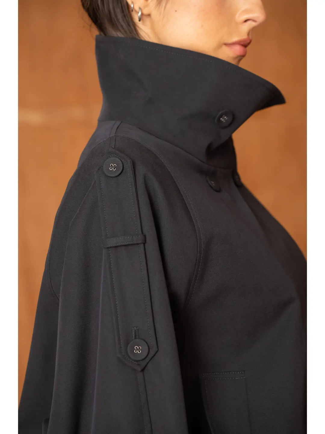 Close-up side view of a person wearing a black high-neck jacket with button details on the shoulder and sleeve, against a warm brown background.