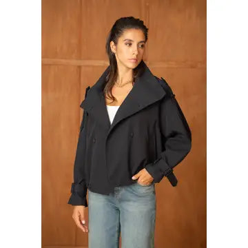 A woman modeling a black double-breasted peplum trench jacket with a large collar and belted cuffs, paired with light blue jeans, set against a warm wood-paneled background.