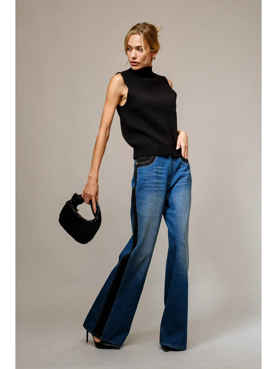 Woman wearing a black sleeveless top and blue jeans, holding a black handbag against a neutral background