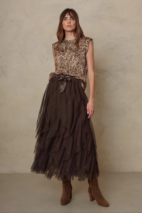 A woman stands against a textured beige studio wall wearing a sleeveless leopard-print top with a waist tie and a long dark brown tiered tulle skirt, paired with brown ankle boots.