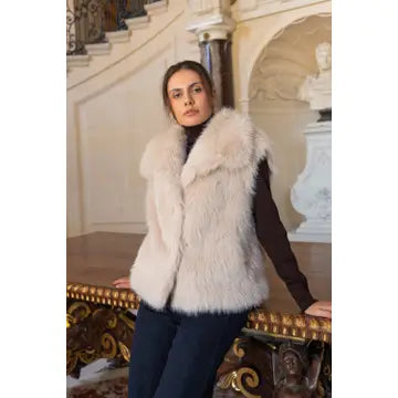 A woman wearing a fluffy cream faux fur vest over a dark brown top and dark pants, posing in an elegant interior setting with a staircase and classical statue in the background.