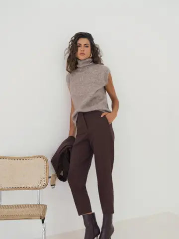 A woman models a sleeveless taupe knit turtleneck sweater and dark brown cropped trousers, standing in a minimalist room with a light-colored wall and a small woven chair.