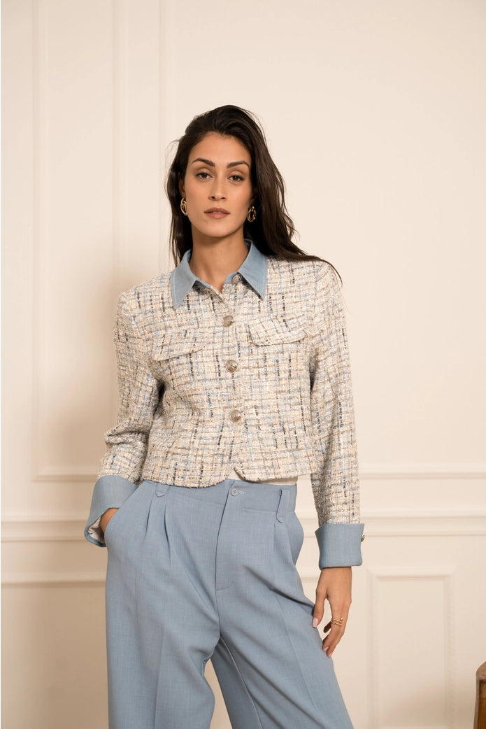 Woman wearing a patterned jacket and light blue pants against a neutral wall.