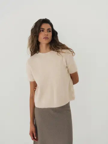 A woman modeling a short-sleeve knit top in a warm off-white/cream color, paired with a textured taupe skirt, against a plain light gray studio background.