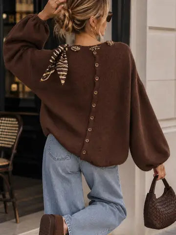 A woman wearing a brown knit sweater with a button-back detail and a striped neck scarf, paired with light blue jeans and holding a brown woven handbag.