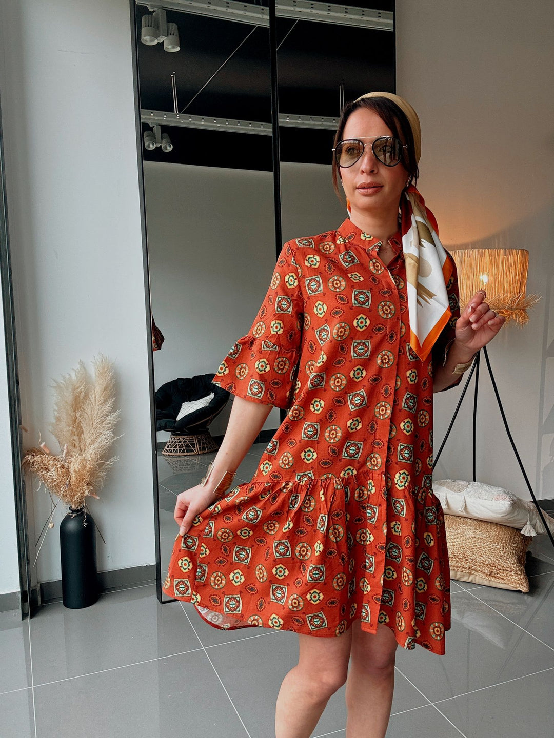 Woman wearing an orange patterned dress holding a clutch in a room.