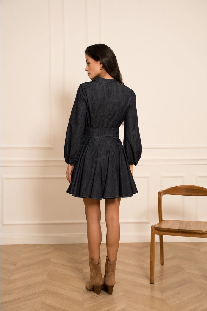 Woman wearing a black dress standing in a room with wooden floor and chair.
