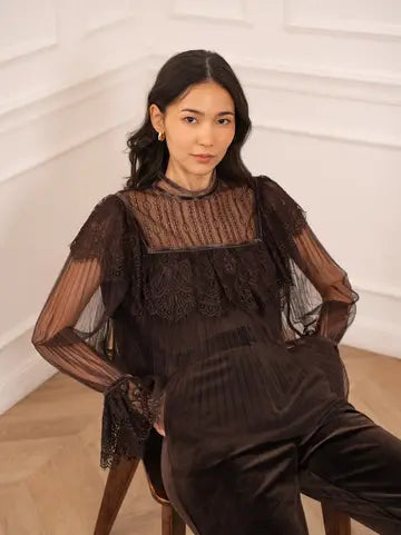 A woman seated on a wooden chair wearing a dark brown sheer lace top with long bell sleeves and matching dark brown velvet pants, posed against a white paneled wall.