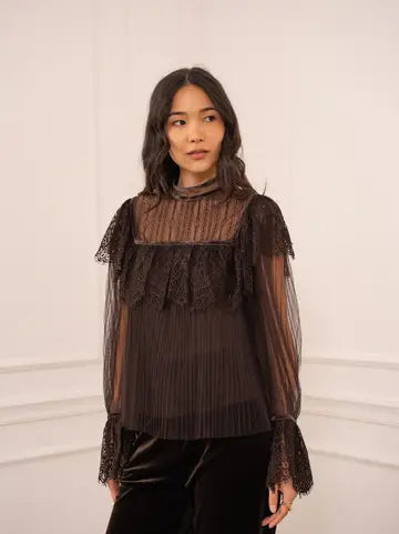 A woman wearing a sheer black long-sleeve blouse with a ruffled yoke and lace cuffs, paired with black pants, standing against a white paneled wall.