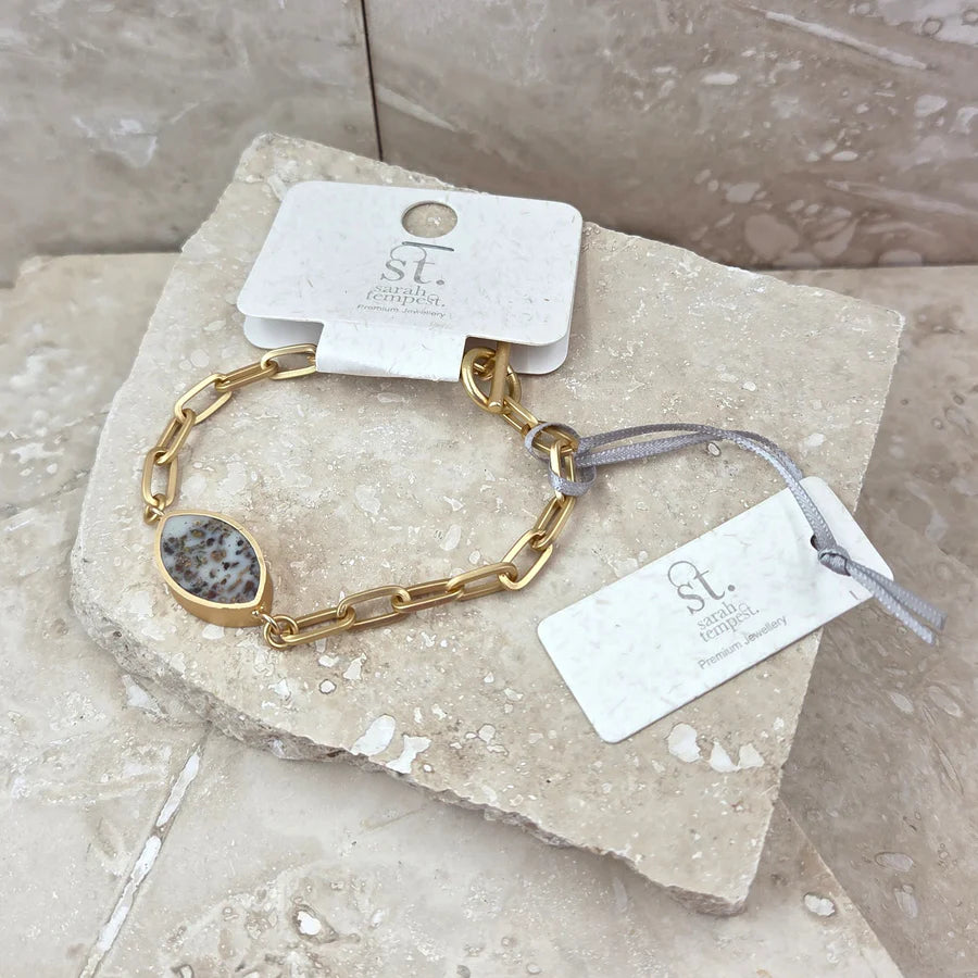 Gold chain bracelet with a stone accent on a marble surface with a brand tag.