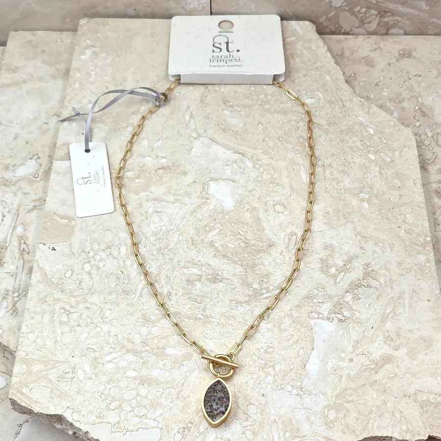 Gold necklace with a pendant on a textured surface, branded 'sarah beth jewelry'.