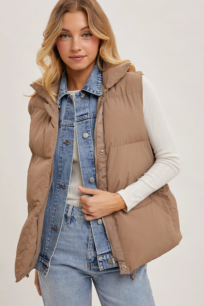 Woman wearing a beige puffer vest over a denim jacket and jeans on a plain background