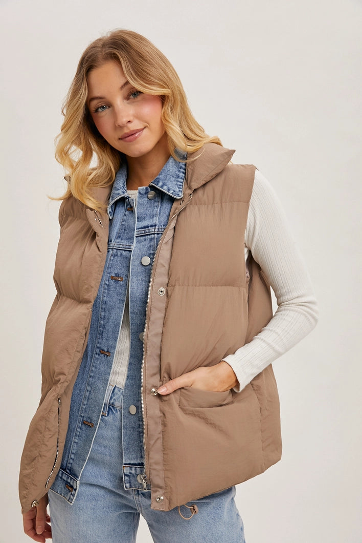 Woman wearing a brown puffer vest over a denim jacket and white shirt on a plain background