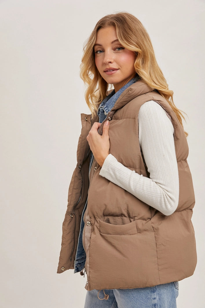Woman wearing a brown puffer vest over a white shirt and blue jeans on a plain background