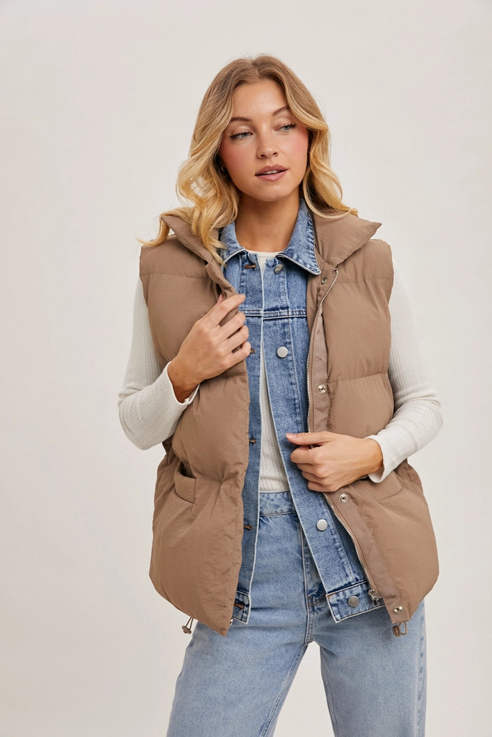 Woman wearing a brown puffer vest over a denim jacket and light blue jeans on a plain background