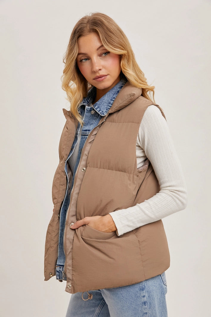 Woman wearing a brown puffer vest over a denim shirt and white sweater on a plain background