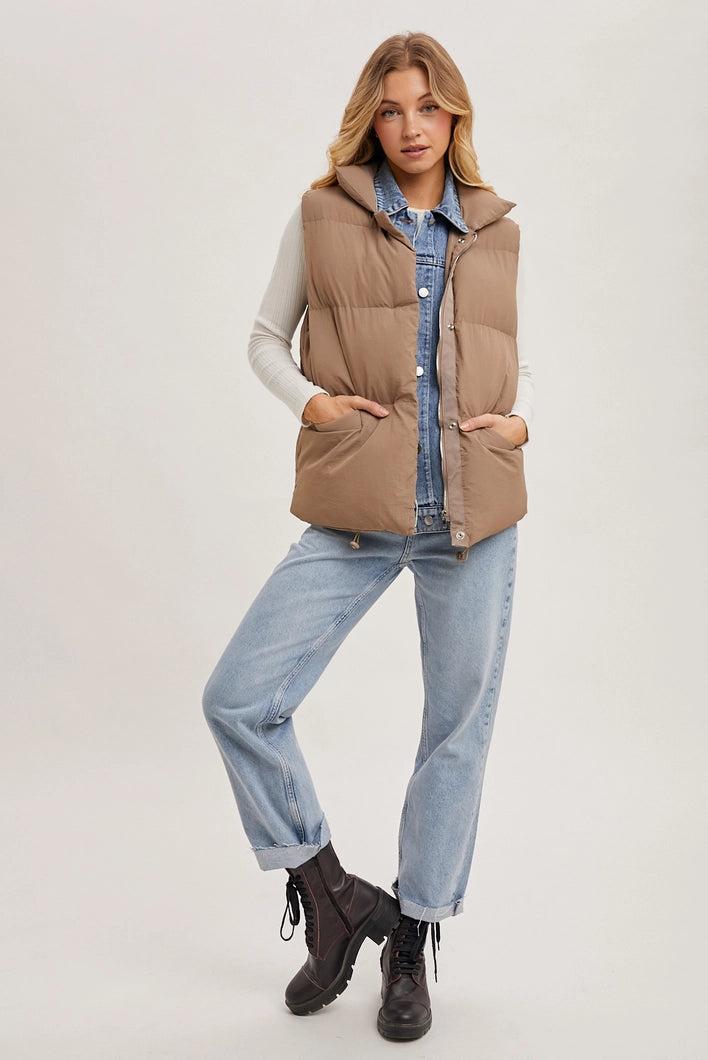 Woman wearing a beige puffer vest over a denim shirt and jeans on a white background