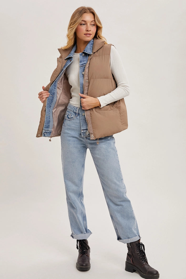 Woman wearing a beige vest over a denim shirt with light blue jeans and black boots on a white background