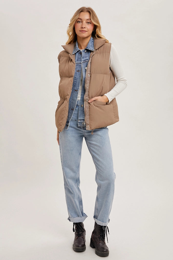 Woman wearing a beige puffer vest over a denim shirt and light blue jeans on a white background
