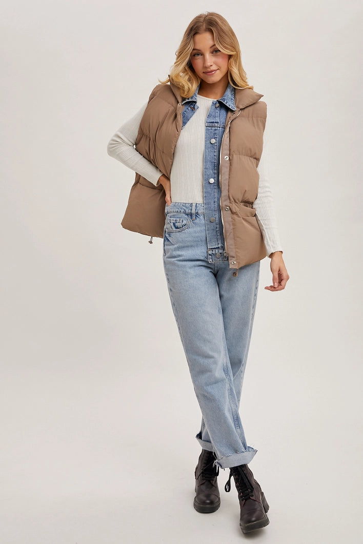 Woman wearing a beige puffer vest over a denim jacket with light blue jeans on a white background