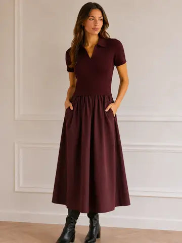 A woman modeling a short-sleeve polo-neck midi dress in deep burgundy with side pockets, paired with black ankle boots, standing against a white paneled wall.