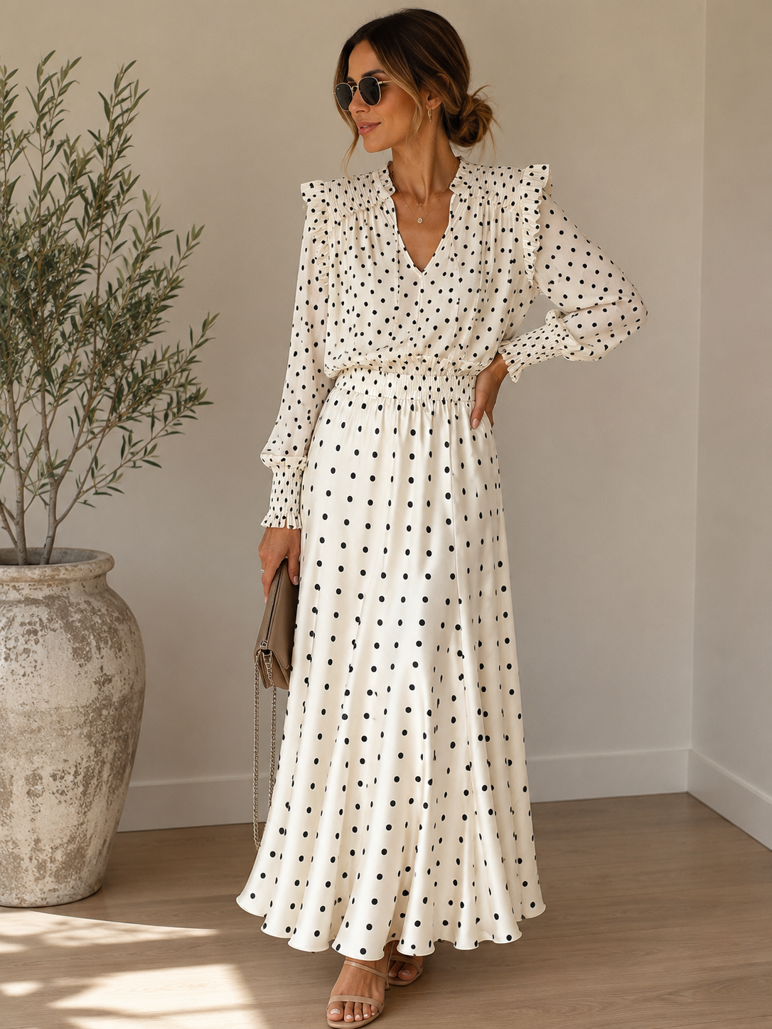 A woman models a long-sleeve white maxi dress with black polka dots, featuring a V-neckline, ruffled shoulders, and a matching tie-waist belt. She is standing in a brightly lit room with a large textured planter on the left.