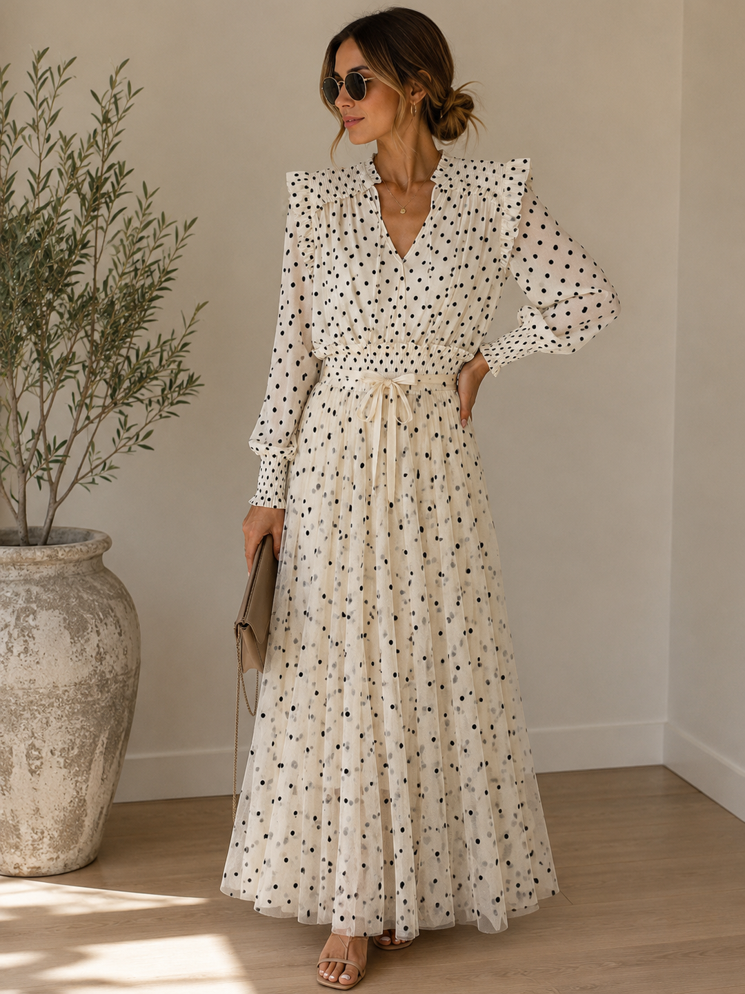 A woman wearing a long-sleeve white maxi dress with a black polka dot pattern, standing in a sunlit room next to a large potted plant.