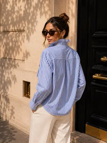 A woman wearing a blue and white vertically striped button-down shirt and cream trousers, photographed from the back in an outdoor urban setting next to a black door.