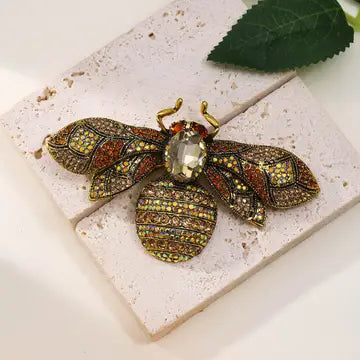 A detailed bee-shaped brooch with gold-tone metal and multicolored rhinestones, displayed on a textured white stone tile with a green leaf in the corner.