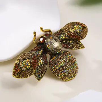 A detailed bee-shaped brooch featuring gold-tone metal, intricate rhinestone patterns in gold, brown, and red, and a clear faceted crystal at the center, resting on a white surface.