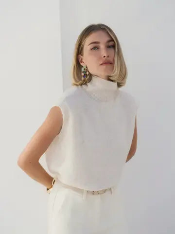 A woman with a blonde bob hairstyle wearing a white sleeveless mock-neck knit top and white trousers, standing against a plain white wall.