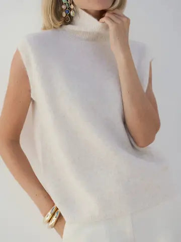 A close-up of a woman wearing a sleeveless, high-neck fuzzy knit top in a warm off-white or cream color, styled with gold jewelry against a plain light background.