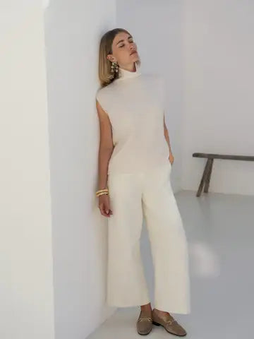 A woman wearing a cream sleeveless mock-neck knit top and matching wide-leg cropped pants, standing in a minimalist white room with a wooden bench in the background.