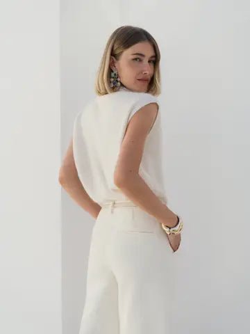 A woman with blonde hair is shown from the back wearing a sleeveless white top and matching white trousers, standing against a plain white wall.