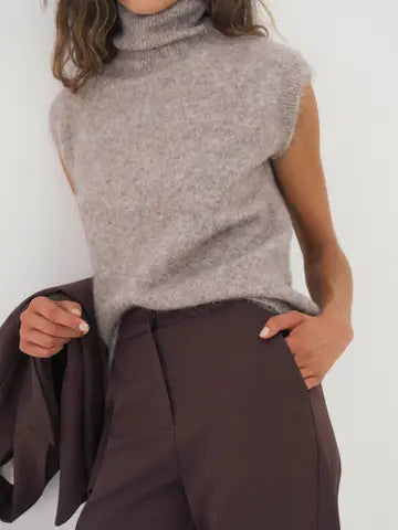 A close-up of a woman wearing a sleeveless taupe-gray turtleneck sweater and dark brown trousers, holding a dark blazer against a plain light background.